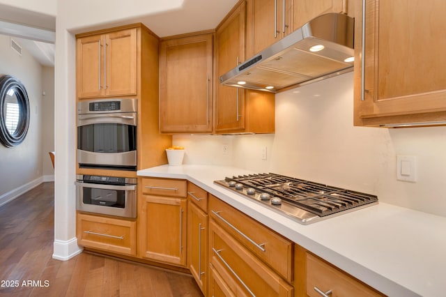 kitchen featuring baseboards, wood finished floors, stainless steel appliances, light countertops, and under cabinet range hood