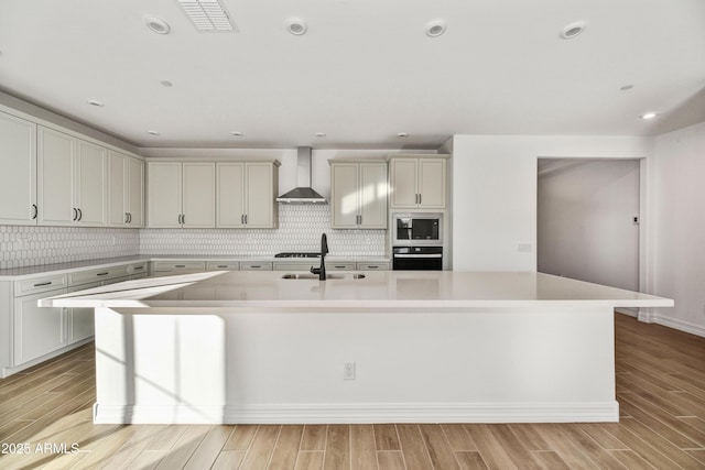 kitchen featuring black oven, built in microwave, sink, a kitchen island with sink, and wall chimney exhaust hood