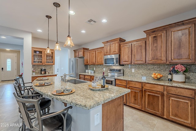 kitchen featuring visible vents, brown cabinetry, appliances with stainless steel finishes, a kitchen island with sink, and a sink