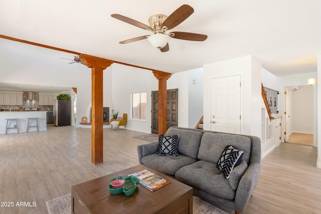 living room featuring ornate columns, a ceiling fan, light wood-type flooring, baseboards, and stairs