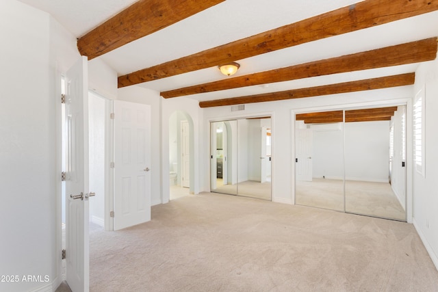 unfurnished bedroom with light carpet, arched walkways, ensuite bath, two closets, and beam ceiling