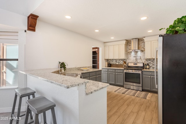 kitchen featuring gray cabinetry, appliances with stainless steel finishes, a sink, wall chimney range hood, and a peninsula