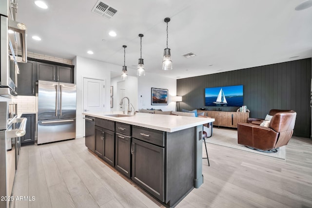 kitchen featuring sink, hanging light fixtures, light wood-type flooring, a kitchen island with sink, and appliances with stainless steel finishes