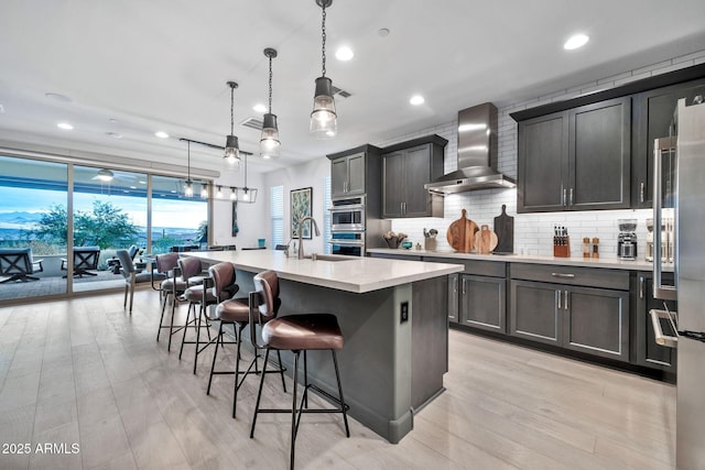 kitchen with high end refrigerator, light hardwood / wood-style floors, hanging light fixtures, a kitchen island with sink, and wall chimney exhaust hood