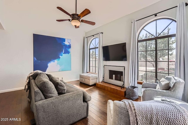 living room featuring a fireplace with raised hearth, wood-type flooring, a ceiling fan, and baseboards