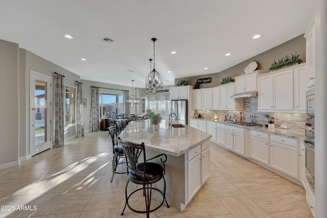 kitchen featuring decorative light fixtures, appliances with stainless steel finishes, light stone countertops, a kitchen island with sink, and white cabinets