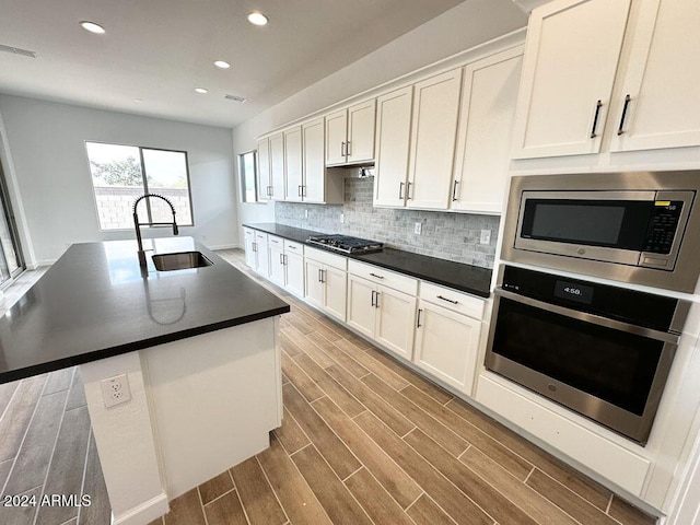 kitchen featuring sink, an island with sink, white cabinets, stainless steel appliances, and backsplash