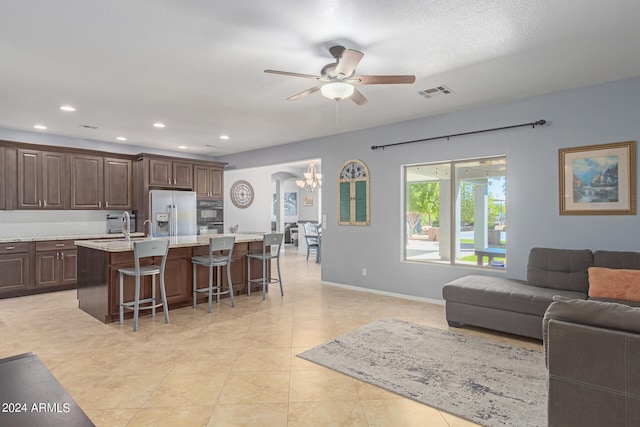 kitchen with a kitchen bar, stainless steel fridge, dark brown cabinets, ceiling fan with notable chandelier, and a center island with sink