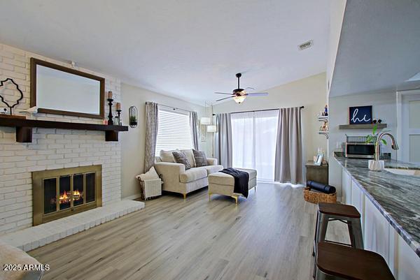 living room with a brick fireplace, wood finished floors, visible vents, and ceiling fan