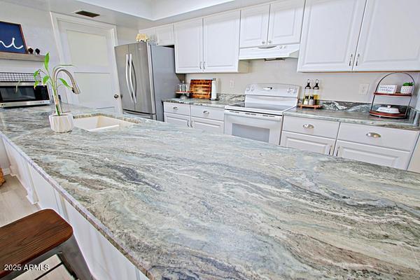 kitchen with light stone counters, under cabinet range hood, freestanding refrigerator, white electric stove, and white cabinets