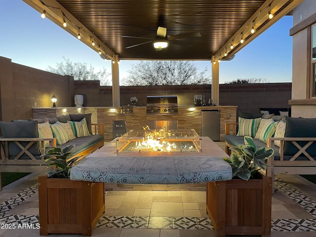 patio terrace at dusk featuring grilling area, ceiling fan, a fire pit, and exterior kitchen