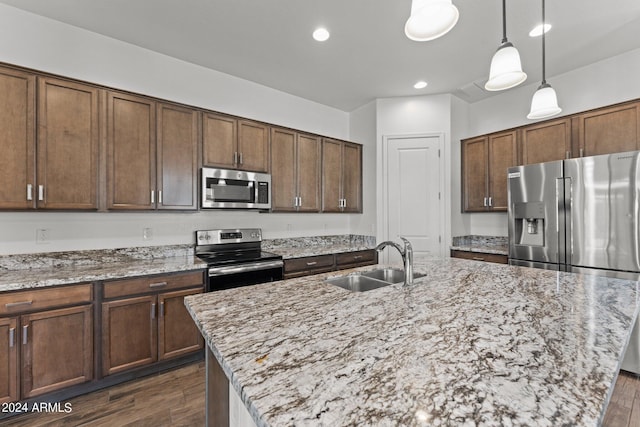 kitchen with dark wood-style floors, appliances with stainless steel finishes, a kitchen island with sink, a sink, and light stone countertops