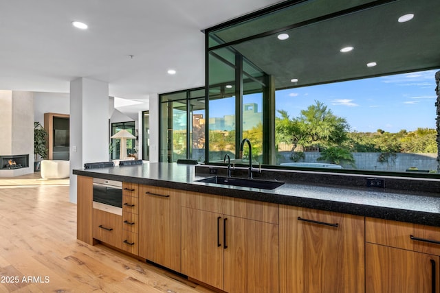 kitchen with a warming drawer, a fireplace, light wood-style flooring, a sink, and dark stone countertops