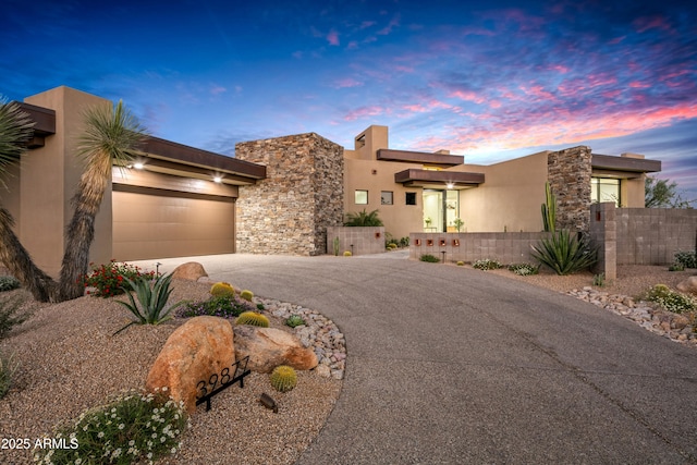 pueblo-style house featuring a garage, stone siding, a fenced front yard, aphalt driveway, and stucco siding