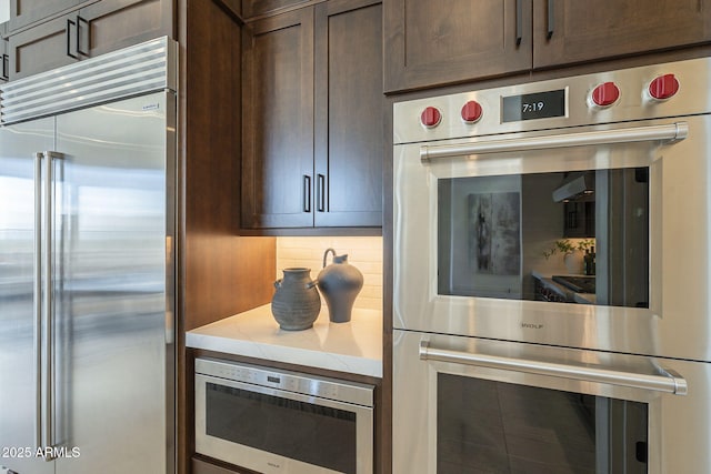 kitchen with dark brown cabinets, built in appliances, decorative backsplash, and light stone counters