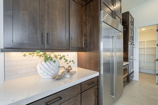 kitchen featuring light stone counters, built in fridge, light tile patterned floors, dark brown cabinetry, and backsplash