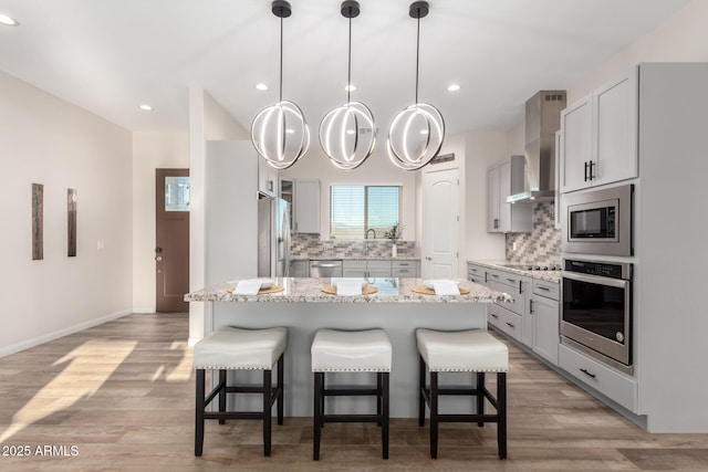 kitchen featuring tasteful backsplash, light stone counters, stainless steel appliances, wall chimney range hood, and a kitchen bar