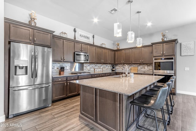 kitchen featuring stainless steel appliances, backsplash, dark brown cabinets, a sink, and light stone countertops