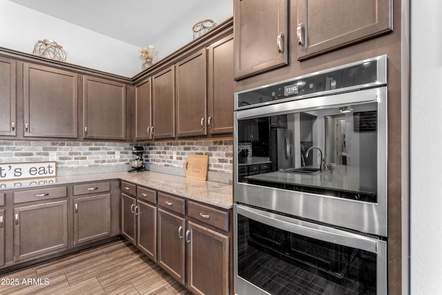 kitchen featuring light stone counters, dark brown cabinets, stainless steel double oven, light wood-style floors, and backsplash