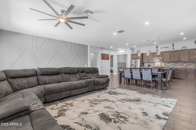 living area featuring a ceiling fan, visible vents, light wood-style floors, and a decorative wall