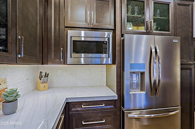 kitchen featuring glass insert cabinets, light stone countertops, dark brown cabinets, stainless steel appliances, and backsplash