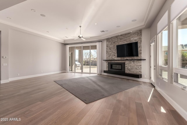 unfurnished living room featuring a stone fireplace, ornamental molding, a raised ceiling, ceiling fan, and hardwood / wood-style floors