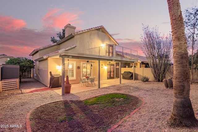 back of house at dusk with a balcony, fence, stucco siding, a tile roof, and a patio area