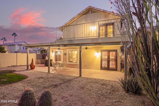 back of house at dusk with stucco siding, a ceiling fan, fence, french doors, and a balcony