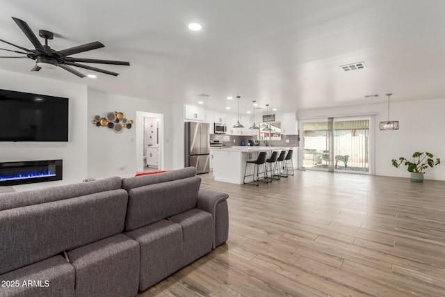 living room featuring light wood finished floors, visible vents, a ceiling fan, a glass covered fireplace, and recessed lighting