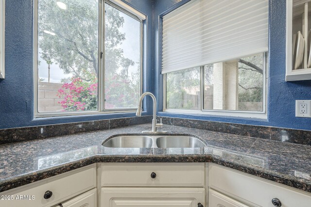kitchen featuring a sink, a textured wall, white cabinetry, glass insert cabinets, and dark stone countertops