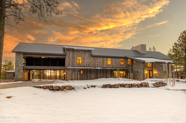 snow covered property with a standing seam roof, a chimney, stone siding, and metal roof