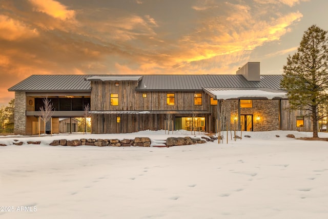 snow covered property with a standing seam roof, a chimney, stone siding, and metal roof