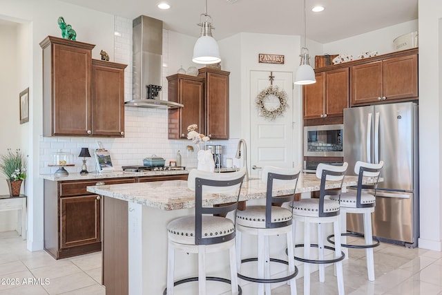 kitchen featuring pendant lighting, appliances with stainless steel finishes, light stone countertops, an island with sink, and wall chimney exhaust hood