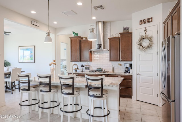 kitchen with appliances with stainless steel finishes, hanging light fixtures, light stone countertops, an island with sink, and wall chimney exhaust hood