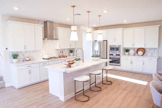 kitchen featuring stainless steel appliances, hanging light fixtures, a kitchen island with sink, white cabinets, and wall chimney exhaust hood