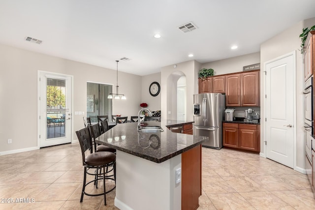 kitchen with an island with sink, stainless steel appliances, sink, dark stone countertops, and pendant lighting