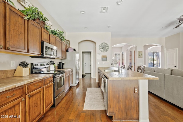 kitchen with appliances with stainless steel finishes, sink, a kitchen island with sink, tile counters, and dark wood-type flooring