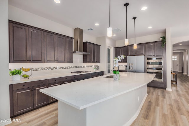 kitchen with arched walkways, a sink, dark brown cabinets, appliances with stainless steel finishes, and wall chimney range hood