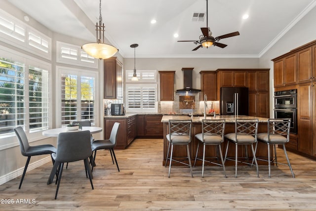 kitchen with multiple ovens, high end black refrigerator, a kitchen island, light stone countertops, and wall chimney range hood