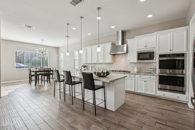 kitchen featuring stainless steel appliances, an island with sink, white cabinetry, and wall chimney exhaust hood