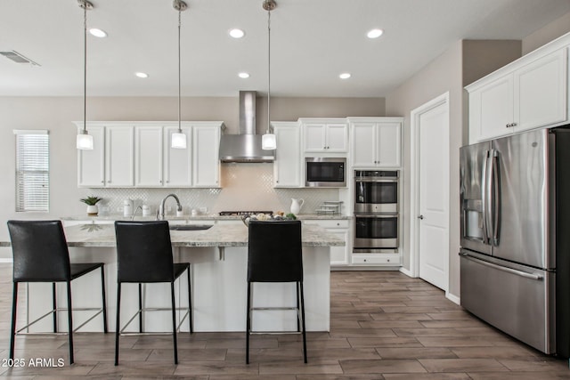 kitchen featuring wall chimney exhaust hood, decorative light fixtures, stainless steel appliances, a kitchen island with sink, and white cabinets