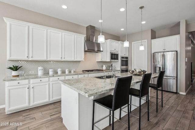 kitchen featuring wall chimney range hood, sink, appliances with stainless steel finishes, an island with sink, and white cabinets