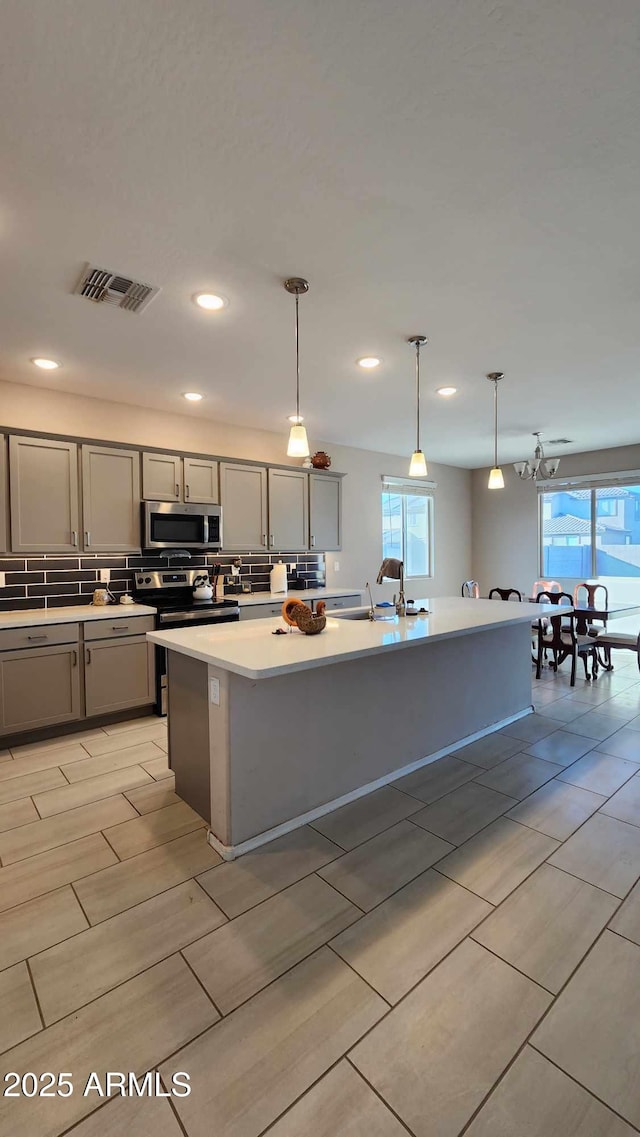 kitchen with gray cabinets, an island with sink, stainless steel appliances, and decorative light fixtures