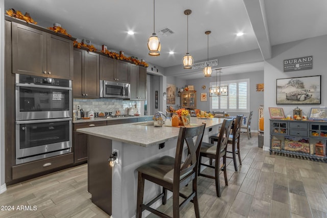 kitchen with stainless steel appliances, light stone counters, hanging light fixtures, dark brown cabinets, and a kitchen island with sink