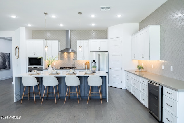 kitchen with an island with sink, stainless steel appliances, decorative light fixtures, wall chimney range hood, and white cabinetry