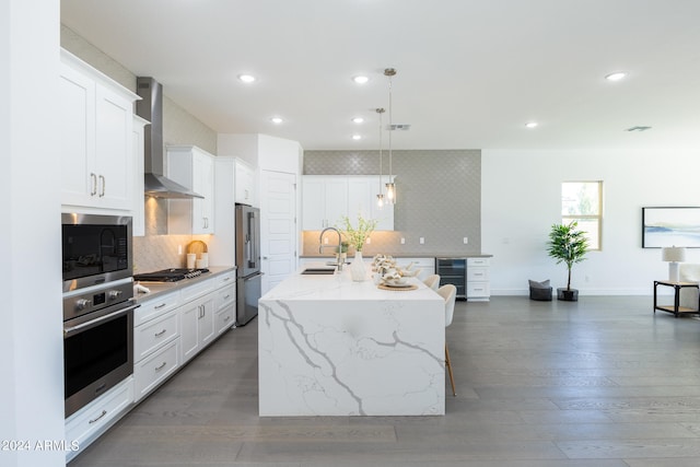 kitchen with a kitchen island with sink, backsplash, stainless steel appliances, wall chimney range hood, and white cabinetry