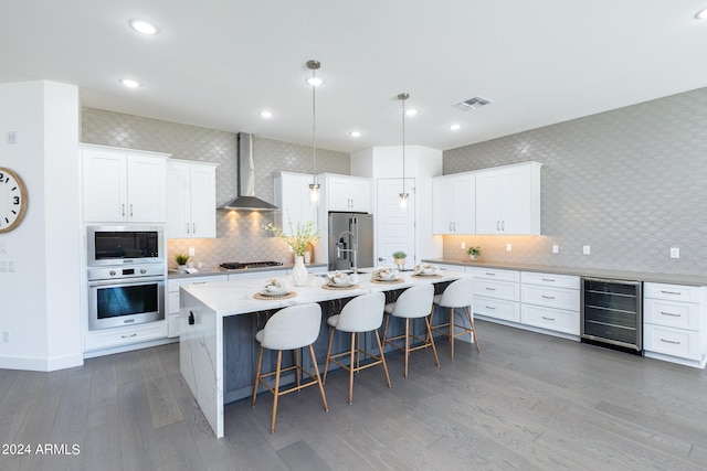 kitchen with white cabinetry, appliances with stainless steel finishes, a center island with sink, beverage cooler, and wall chimney range hood