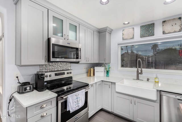 kitchen with gray cabinetry, a sink, appliances with stainless steel finishes, light countertops, and glass insert cabinets