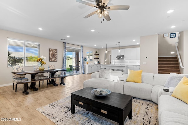 living room featuring light wood-style flooring, ceiling fan, baseboards, recessed lighting, and stairs