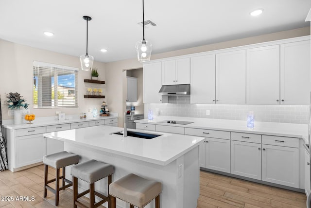 kitchen with light wood-style flooring, a sink, under cabinet range hood, black electric cooktop, and a center island with sink